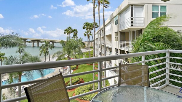 Ein Balkon mit Glastisch und Stühlen, der einen Blick auf ein Schwimmbad und Palmen bietet, mit einem Gebäude im Hintergrund.