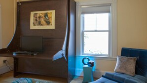 Une pièce avec un bureau incurvé, une télévision, une fenêtre, un canapé bleu et une petite table avec un haut-parleur.