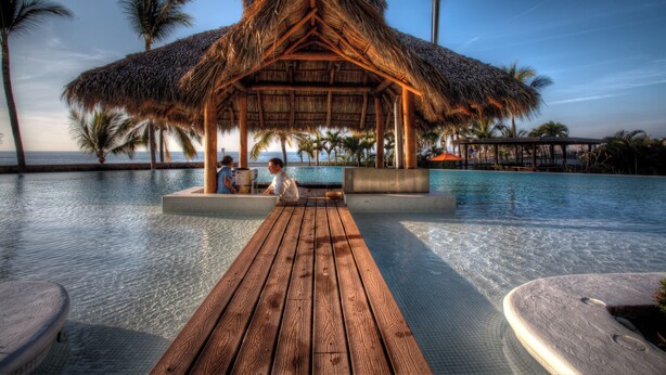 A wooden deck leading to a pool area with a thatched-roof structure, surrounded by palm trees and a clear sky. - Molino de Agua Next to Los Muertos Beach! 3BD Condo for rent in Los Muertos Beac (Puerto Vallarta)