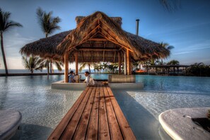 A wooden deck leading to a pool area with a thatched-roof structure, surrounded by palm trees and a clear sky. - Molino de Agua Next to Los Muertos Beach! 3BD Condo for rent in Los Muertos Beac (Puerto Vallarta)