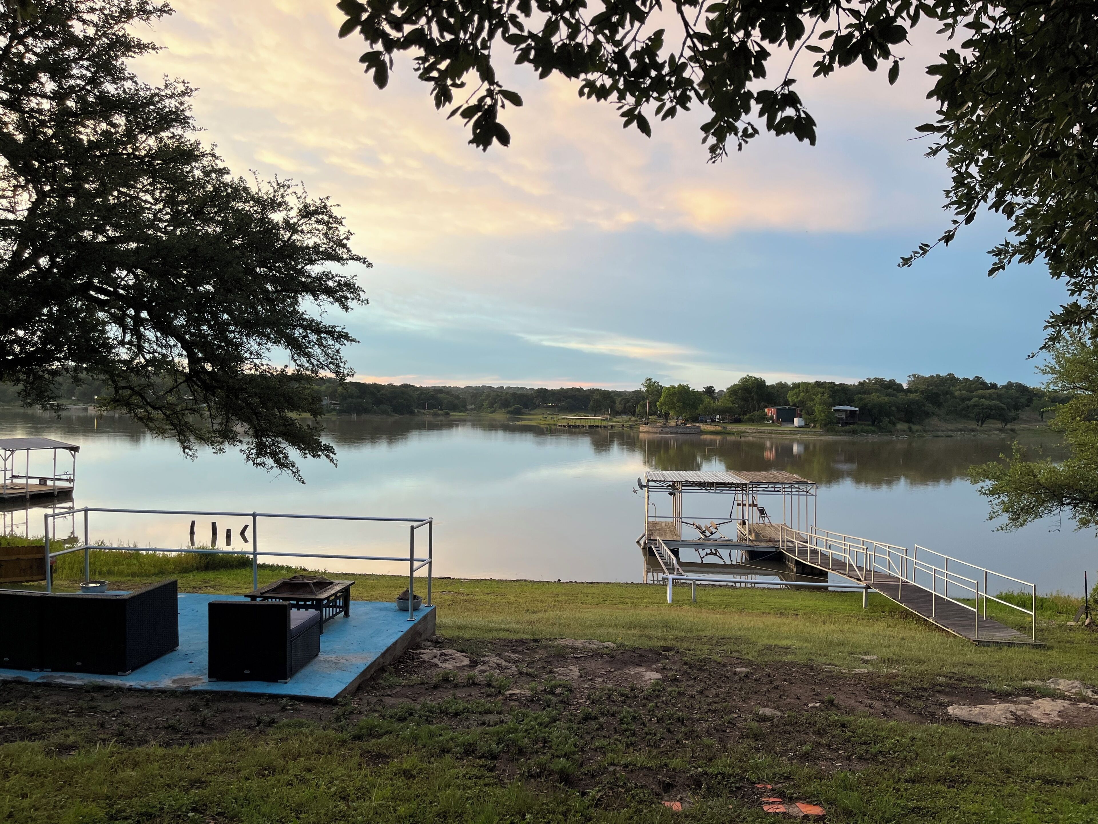 A lakeside view with a dock, a covered seating area, and a grassy field.