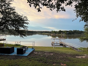 A lakeside view with a dock, a covered seating area, and a grassy field.