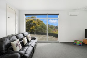 A room with a black leather sofa, a television, and a large window with a view of trees and houses.