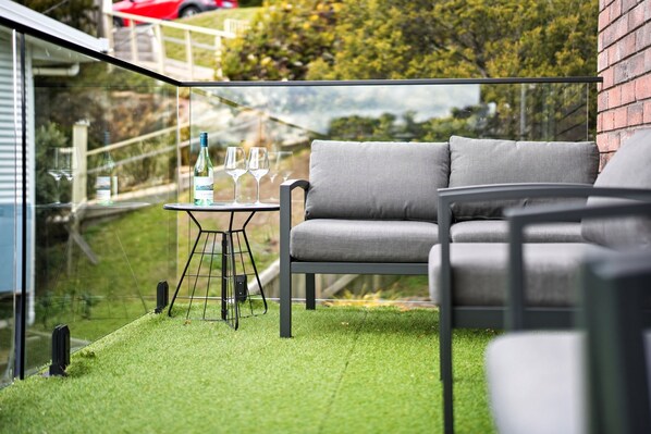 A balcony with a grey sofa, a small table with glasses and a bottle, and a glass railing. - Close to city, tranquil setting (West Hobart)