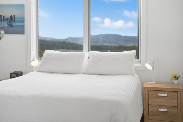 A bedroom with a bed, two bedside lamps, a nightstand with a lamp, and a window with a view of mountains.