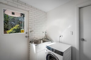 A compact laundry room with a washing machine, a sink, and a window providing a view of greenery outside.