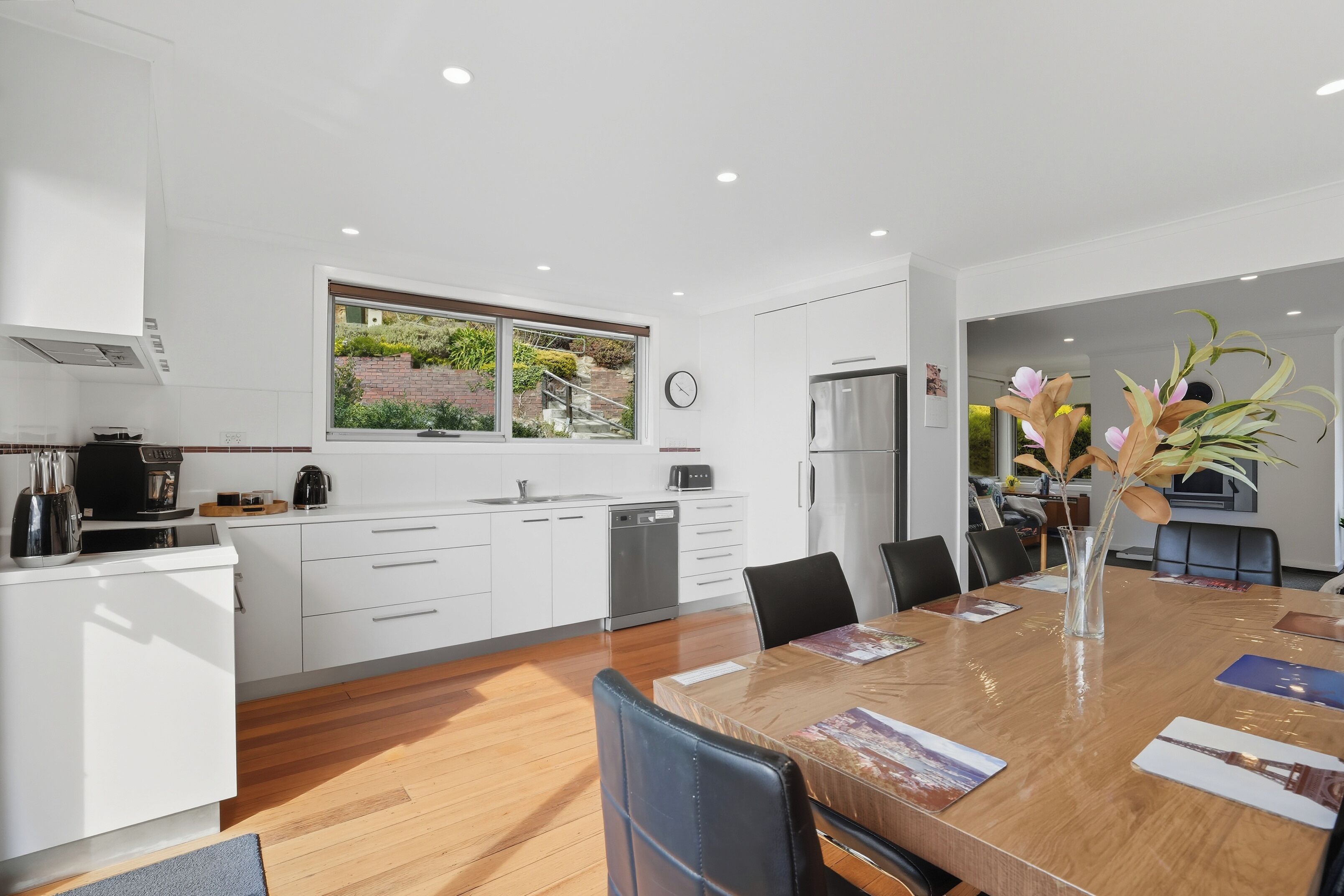 A modern kitchen with white cabinets, a stainless steel refrigerator, and a window overlooking a garden.