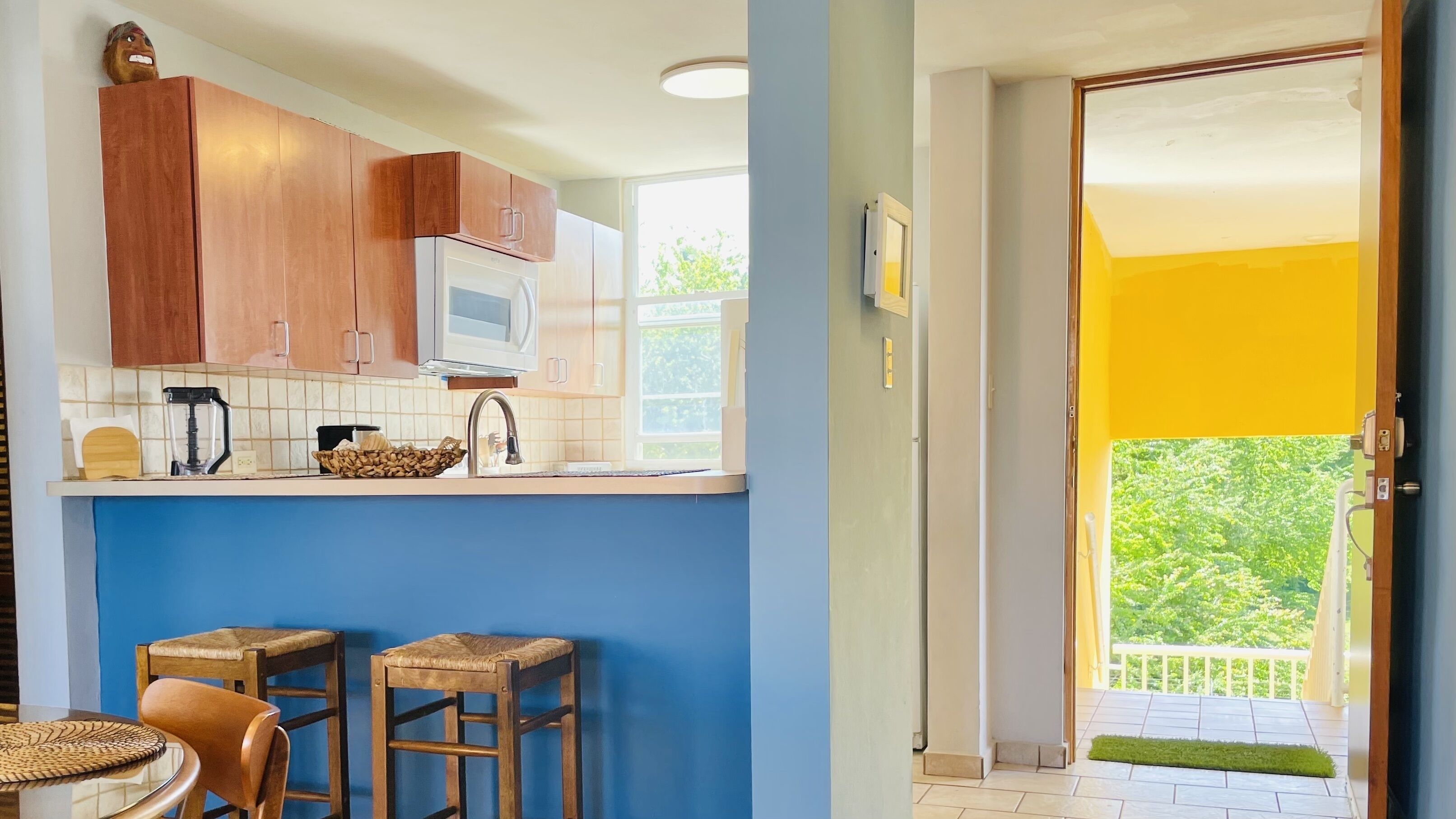 A kitchen with wooden cabinets, a microwave, and a sink. There are two stools and a table with a woven placemat. A balcony with a yellow door is visible.