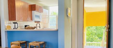 A kitchen with wooden cabinets, a microwave, and a sink. There are two stools and a table with a woven placemat. A balcony with a yellow door is visible.