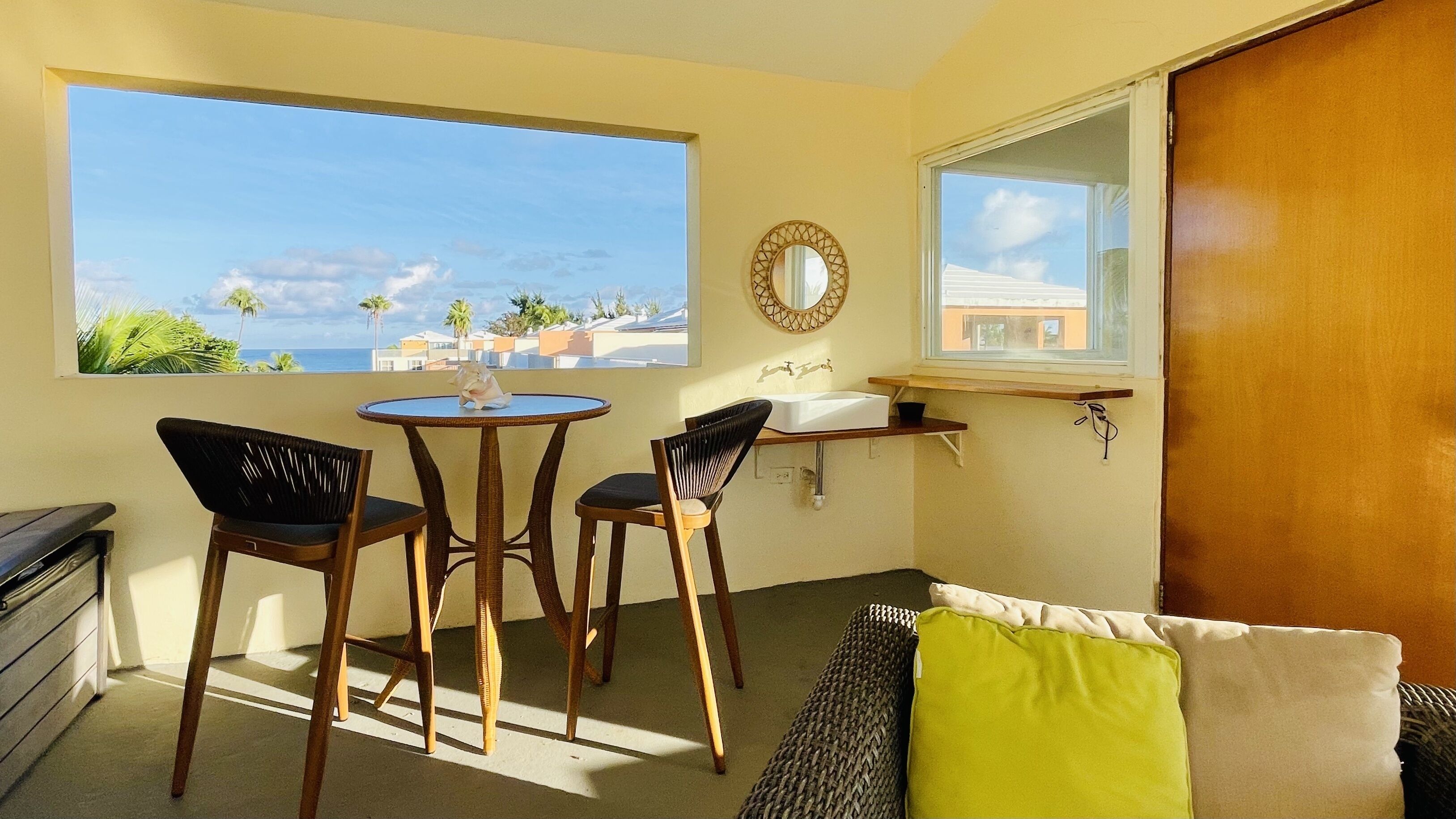 A room with a large window, a round table, two chairs, a small sink, a mirror, and a wooden door.