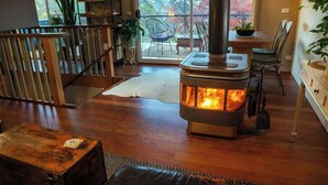 A cozy room with a wood-burning stove, a dining area with chairs, and a view through a glass door. - TreeTops House near Bright with Mtn & pool views (Tawonga South)