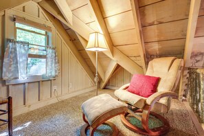 A cozy attic room with a wooden ceiling, a window with sheer curtains, a rocking chair with a red pillow, and a small ottoman.