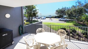 A balcony with a table and chairs, a grill, and a view of a parking lot and greenery.