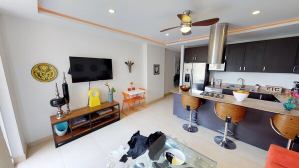 A modern kitchen with dark cabinets, a stainless steel range hood, and a built-in oven. There is a dining area with a white table and orange chairs, and a TV mounted on the wall.