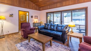 A cozy living room with a leather sofa, wooden coffee table, and a large window offering a view of the outdoors. - Cozy Pet Friendly Cabin, Wood Fireplace, Hot Tub, SHARC passes, close to Village (Sunriver)
