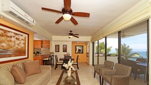 A spacious living room with a sofa, armchairs, a coffee table, and a dining area with chairs and a table. There are ceiling fans, a large window with a view, and an air conditioning unit. - 😍ROMANTIC GETAWAY😍AMAZING OCEAN VIEWS -HOP, SKIP & JUMP TO THE BEACH -GR8 POOL (Puerto Vallarta)