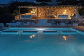 Zona junto a la piscina con muro de piedra, mobiliario exterior y una pérgola.