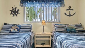 A bedroom with two beds, a nightstand, a lamp, and nautical-themed decor. - Beachy Madeira Getaway | Tropic Breeze Unit 18 – Steps to Beach, Pool Access (Madeira Beach)