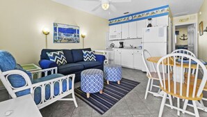 A compact living space with a sofa, ottomans, a dining table with chairs, a ceiling fan, a kitchen area with cabinets and appliances, and a painting on the wall. - Beachy Madeira Getaway | Tropic Breeze Unit 18 – Steps to Beach, Pool Access (Madeira Beach)