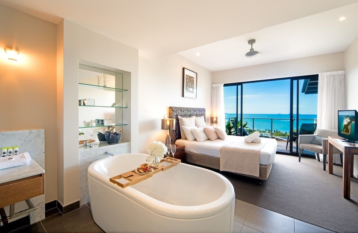 A modern hotel room with a large bed, a bathtub, a balcony with a sea view, and a television.