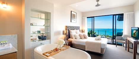 A modern hotel room with a large bed, a bathtub, a balcony with a sea view, and a television.