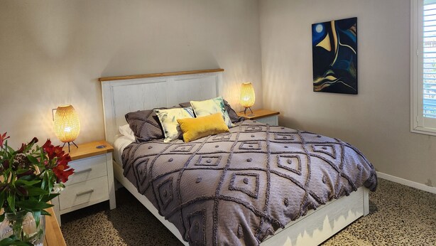 Een slaapkamer met een bed, twee bedlampen, een nachtkastje, een schilderij en een raam met jaloezieën.