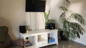 A living room with a flat-screen TV, a white entertainment unit with speakers, a brown sofa, a large potted plant, and a dark rug.