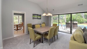 Une salle à manger spacieuse, dotée d’une grande table et de chaises jaunes, d’un lustre moderne et offrant une vue sur une piscine et un jardin grâce à de grandes fenêtres.