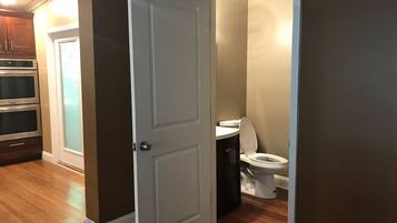Une pièce avec un plancher en bois, une porte blanche, des toilettes blanches et une petite fenêtre avec des stores.