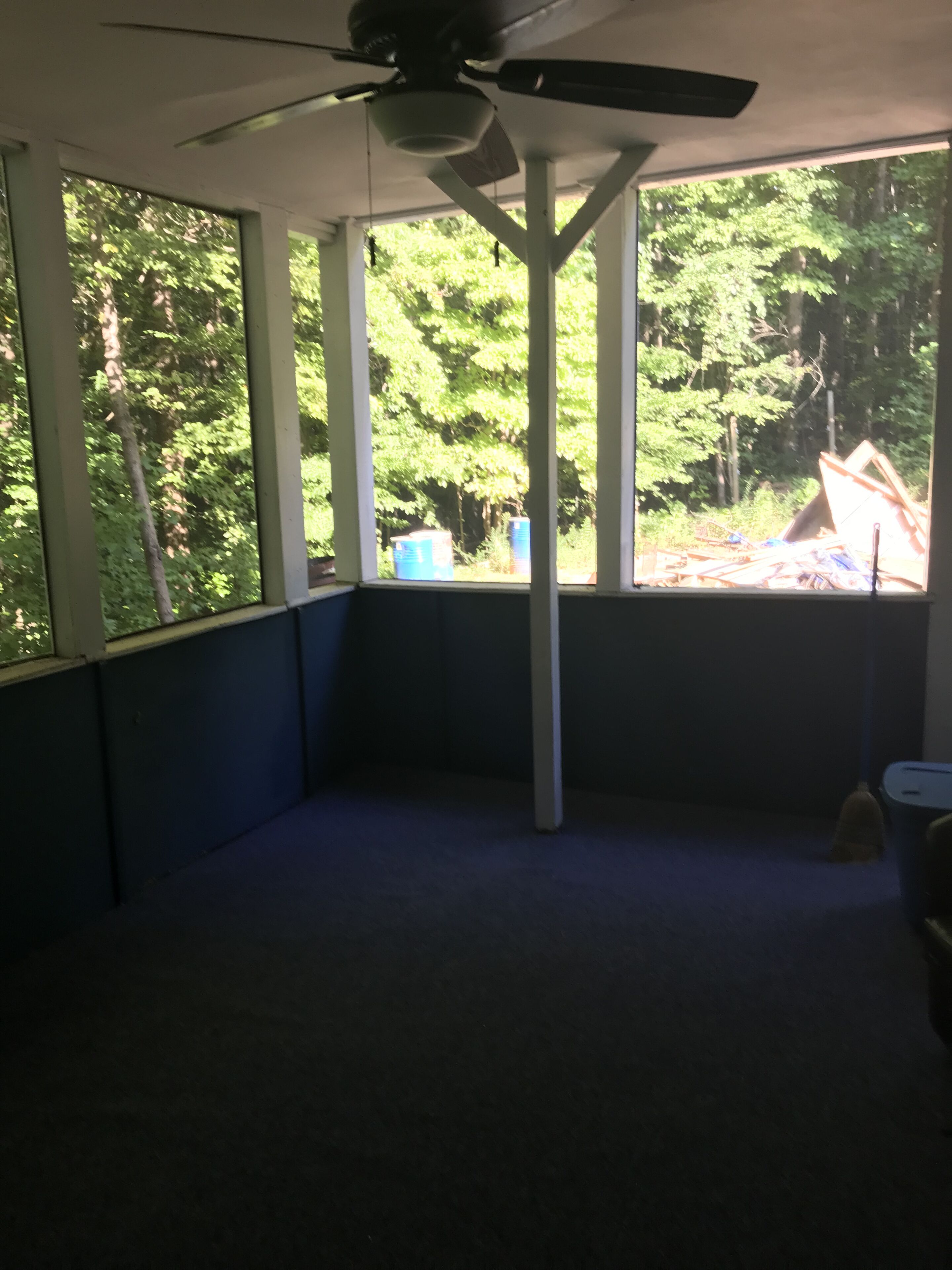 A room with a ceiling fan, large windows, and a view of trees and outdoor debris.