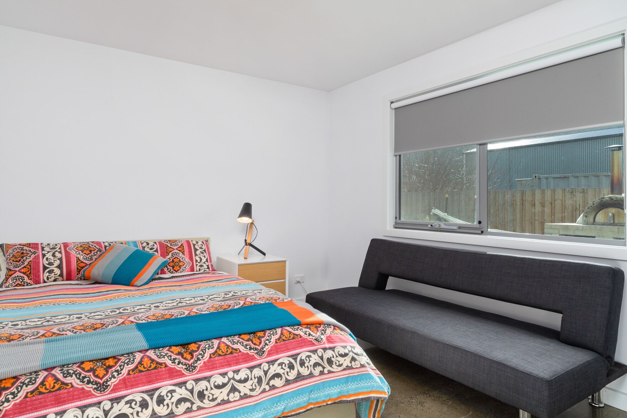 Une chambre à coucher avec un lit, une table de chevet, une lampe, un canapé gris et une fenêtre donnant sur l’extérieur.