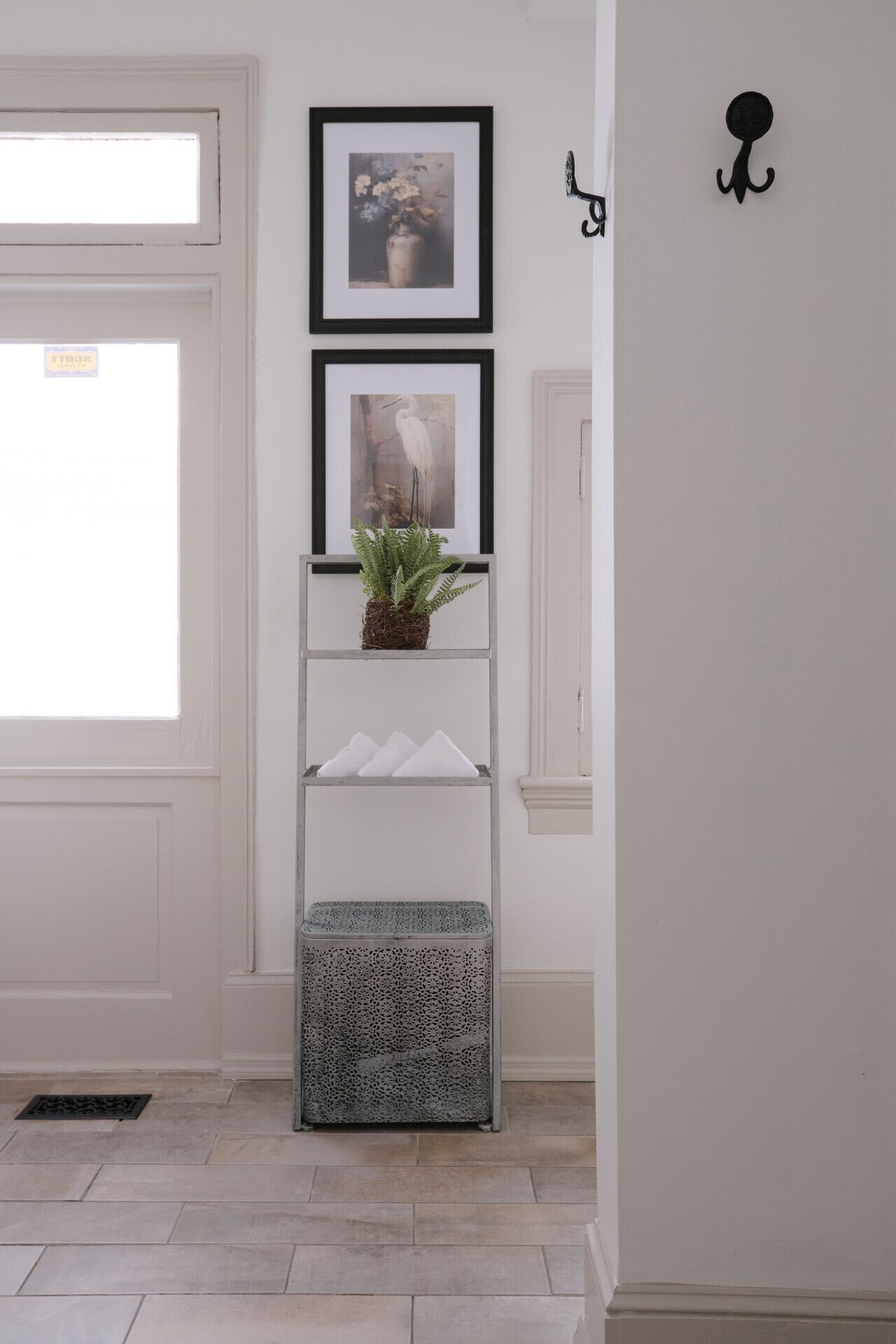 A hallway with a white door, a framed picture of flowers, a framed picture of a bird, a small plant on a shelf, and a decorative wall hook.