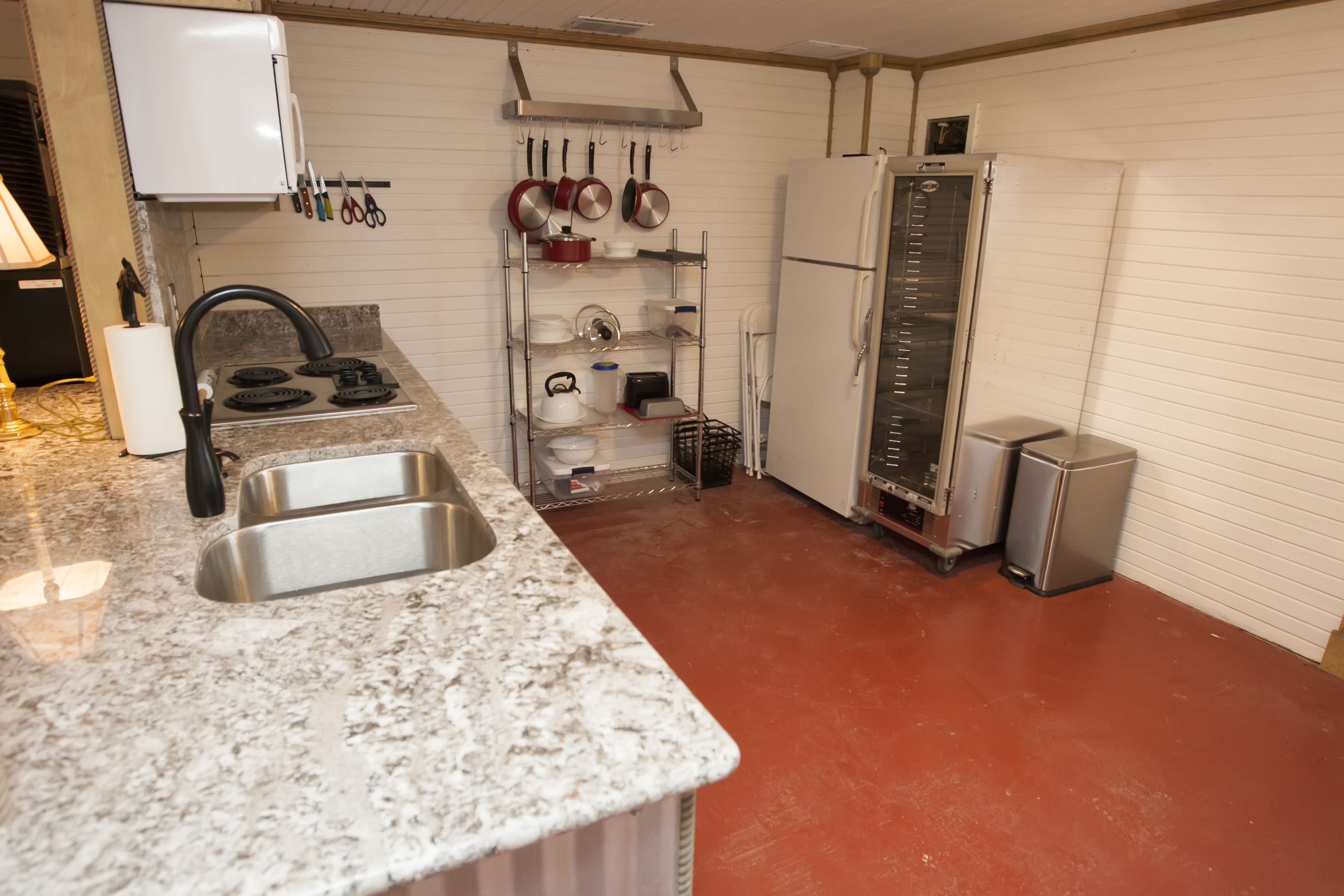 Una cocina con encimera de granito, pileta de acero inoxidable, alacenas blancas y piso rojo.