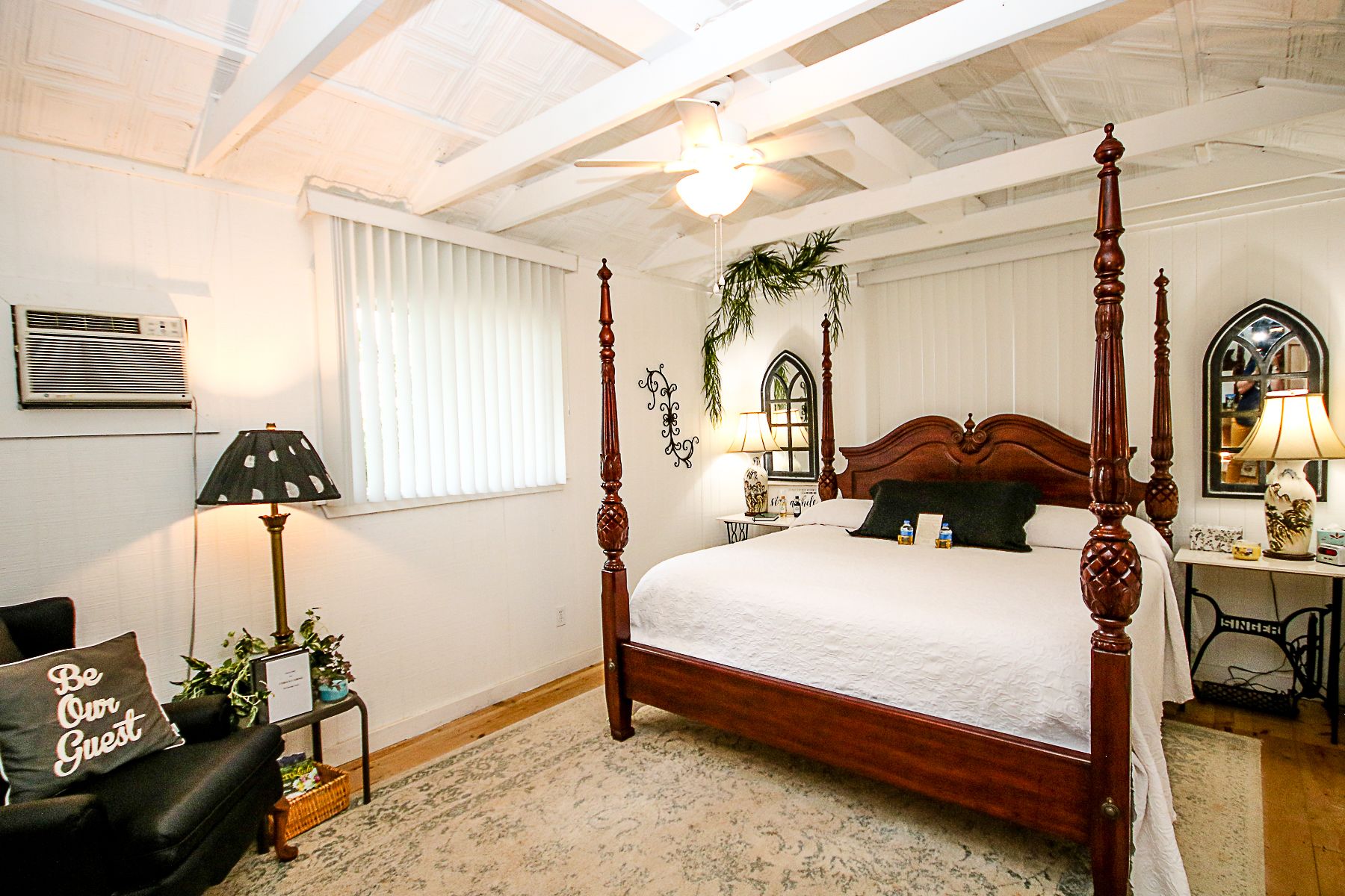 Una cama con dosel y tablero de madera, una mesita de noche con lámpara y una silla con almohada que dice "Be Our Guest".