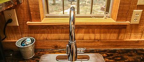 A stainless steel kitchen sink with a window above it, wooden walls, and a small metal bucket on the counter.
