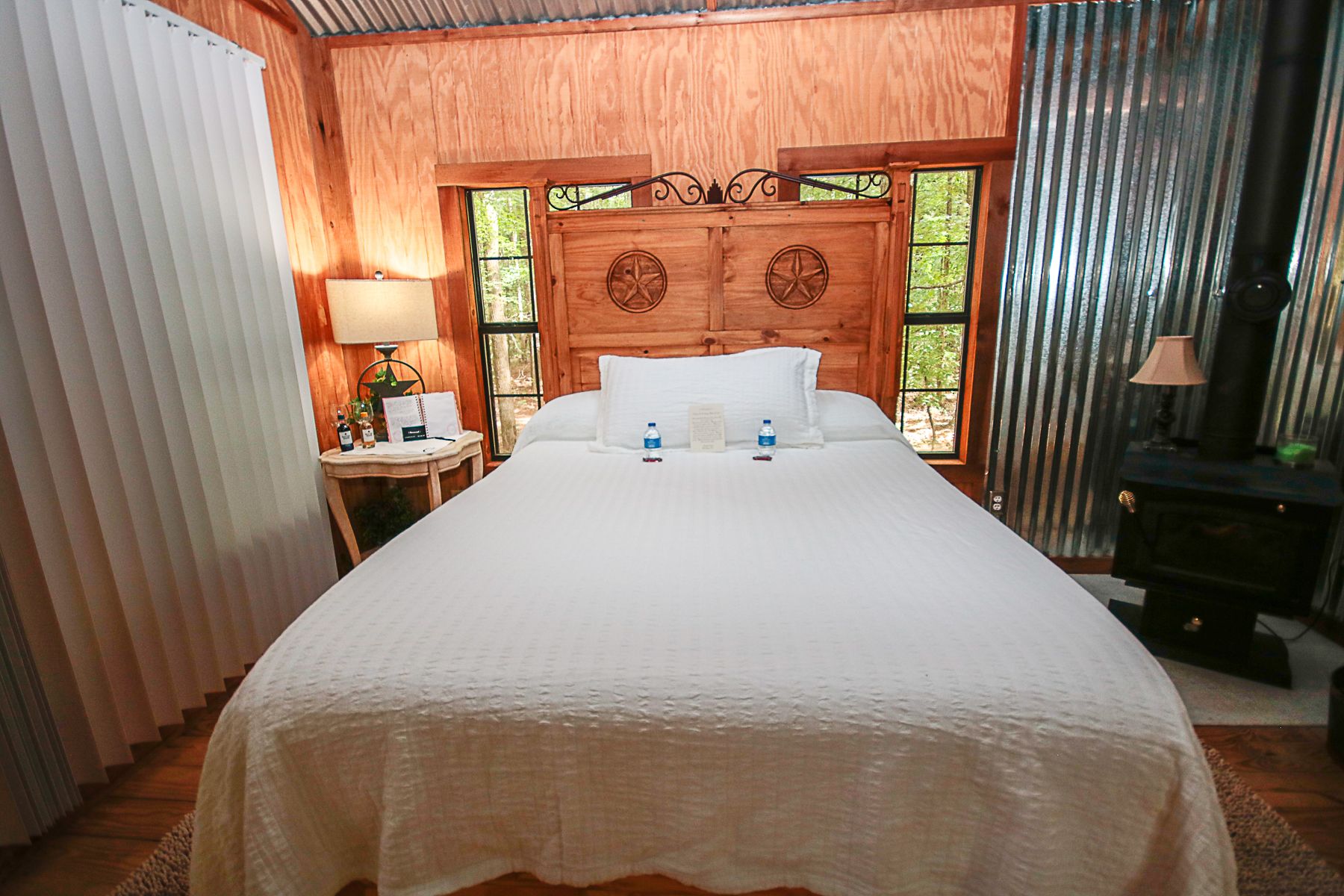 A hotel room with a large bed, wooden headboard, a nightstand with a lamp, and a view of trees through the window.