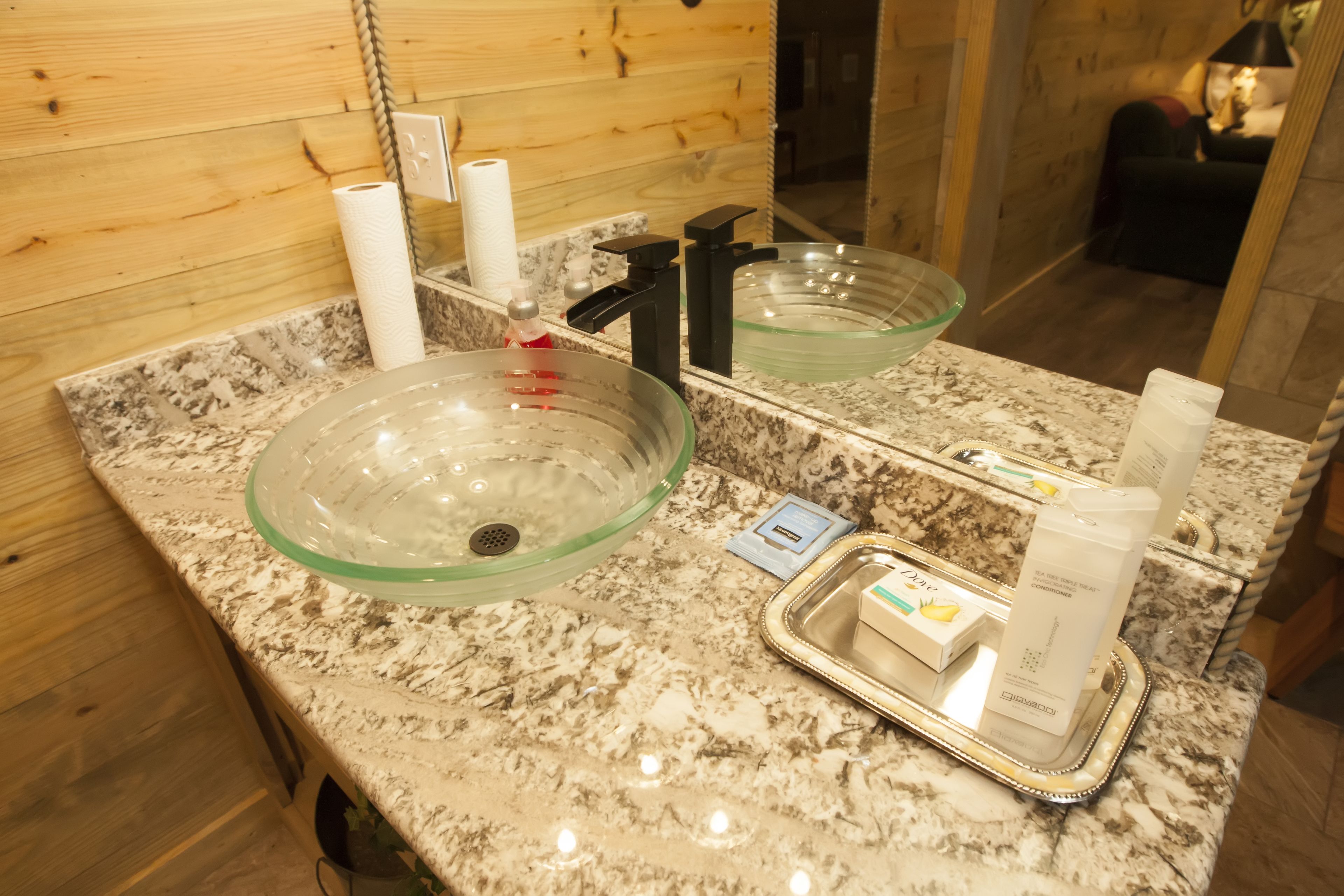 A bathroom with a granite countertop, a glass bowl sink, a mirror, and toiletries on a tray.