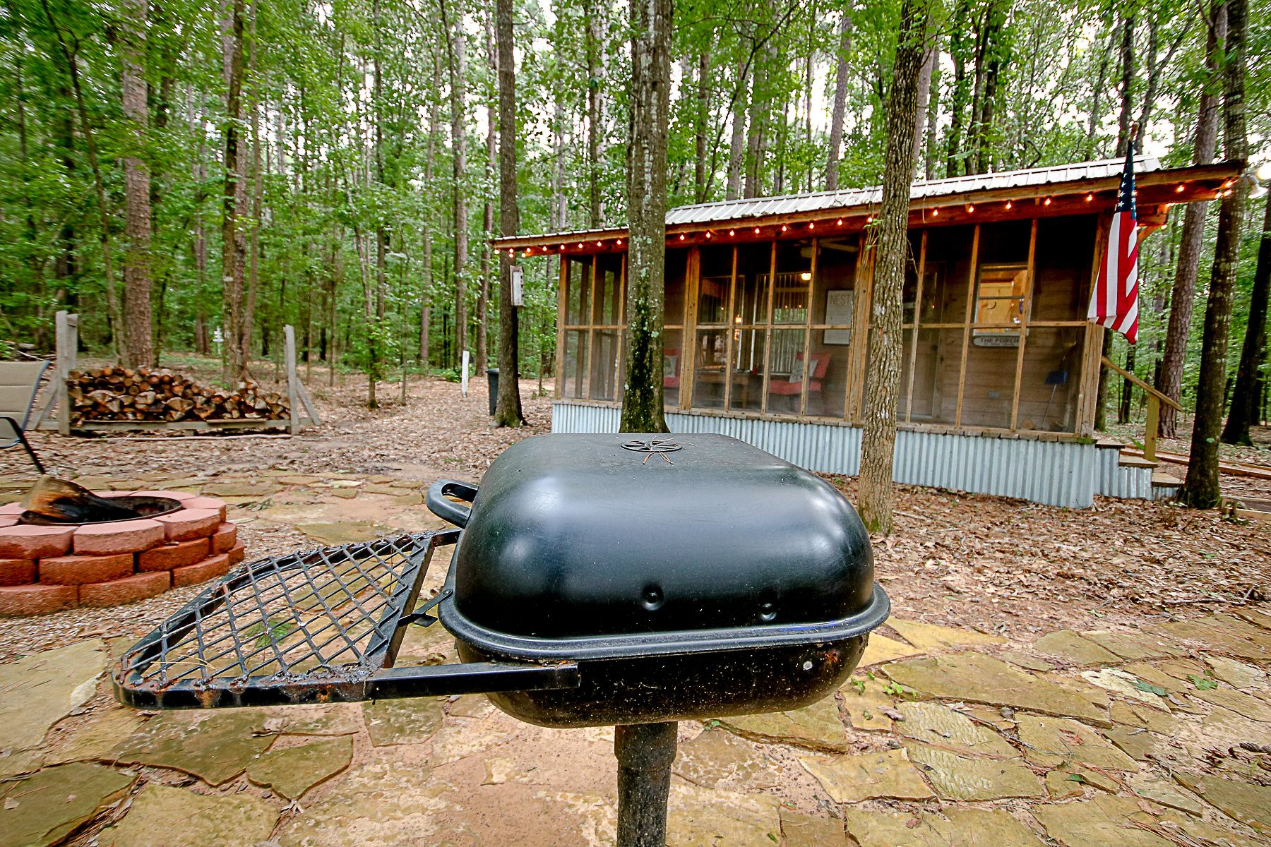 A rustic outdoor grill setup in a wooded area with a small wooden structure in the background.
