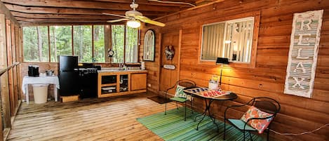 Innenansicht einer Holzhütte mit Küche, Essbereich und großem Fenster mit Blick auf einen Wald.