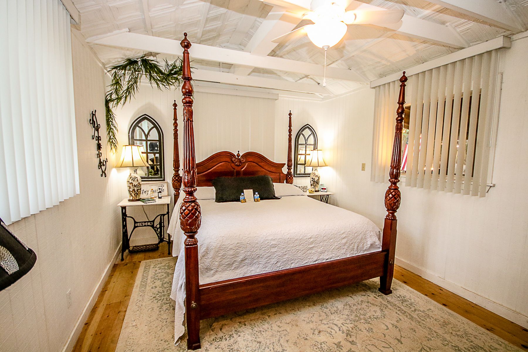 A four-poster bed with a wooden headboard, a nightstand with a lamp, a small table with a vase, and a window with blinds.