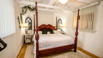 A four-poster bed with a wooden headboard, a nightstand with a lamp, a small table with a vase, and a window with blinds.