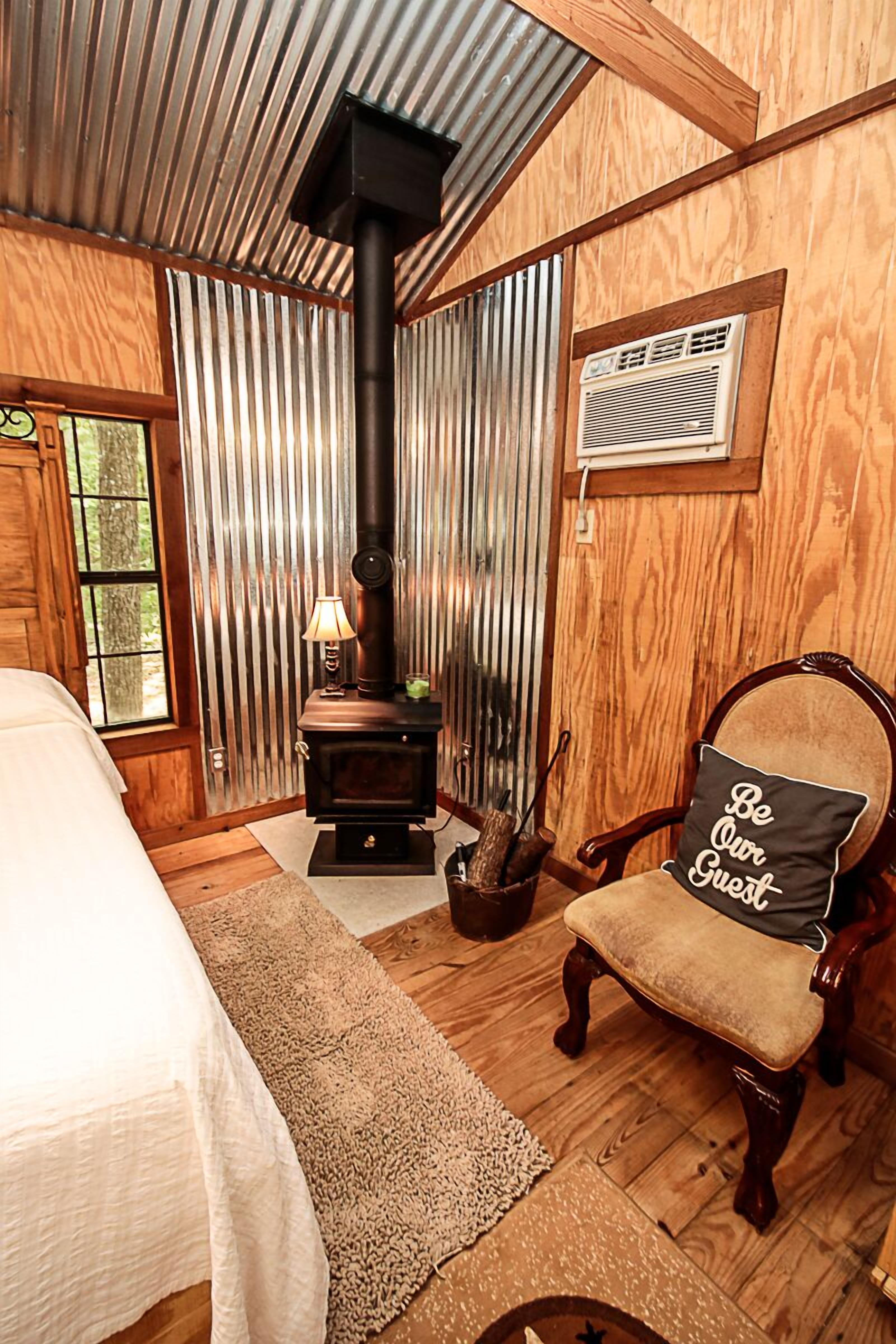 A cozy room with a wood-burning stove, a bed, a chair with a cushion, and a window with curtains.