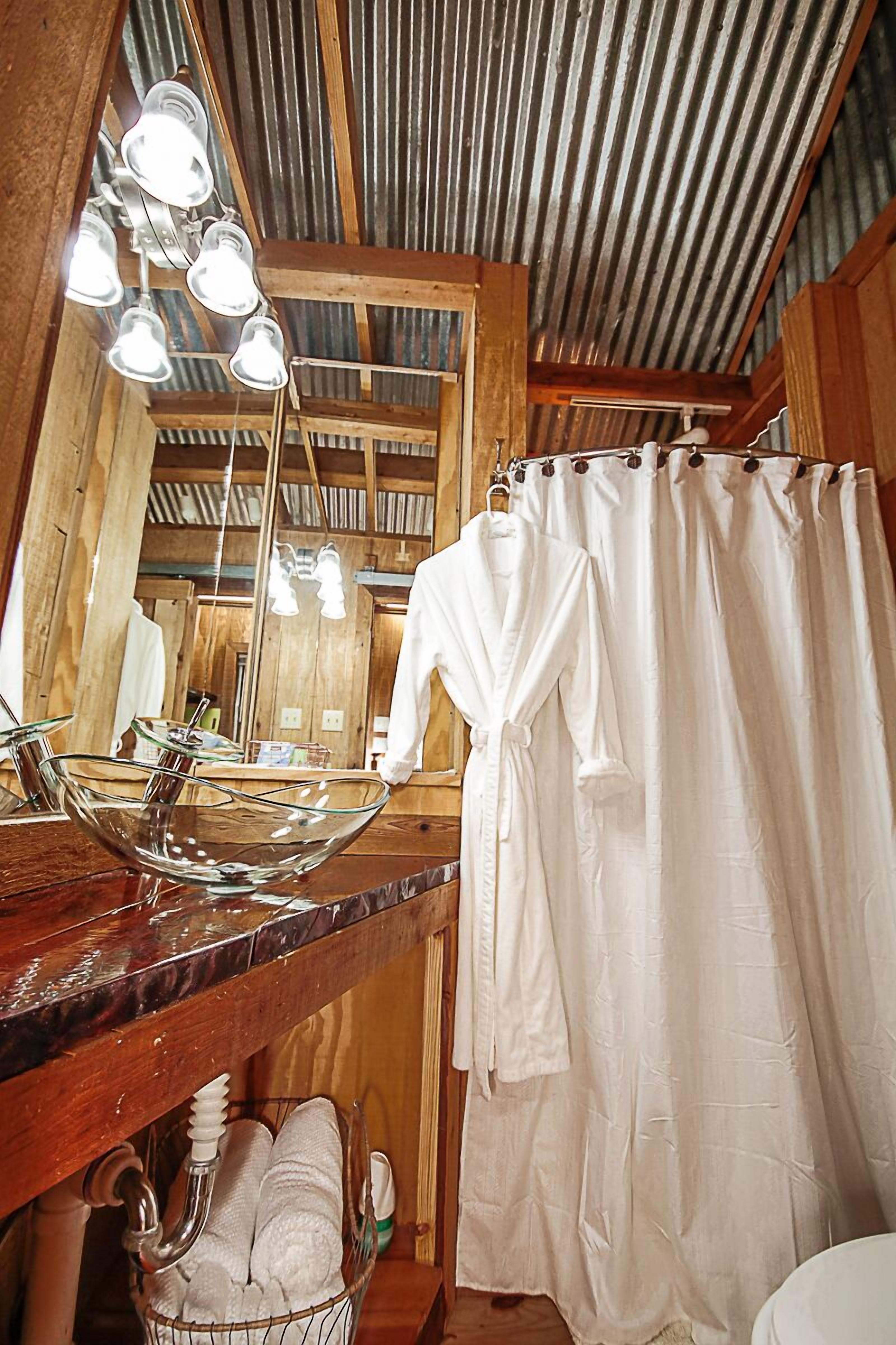 Une salle de bain avec un plafond en bois, un grand miroir, un lavabo en verre et un peignoir blanc accroché au mur.