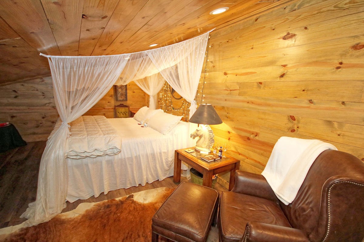 Un dormitorio de cabaña de madera con una cama con dosel, una mesita de noche, una lámpara y un sillón de cuero.
