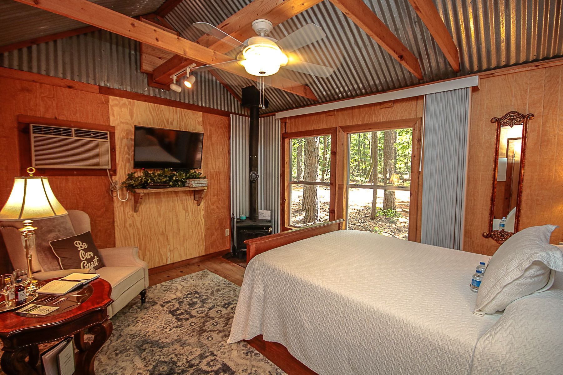 A cozy bedroom with a bed, a desk with a lamp, a chair, a television, and a window with a view of trees.