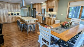 A spacious kitchen with wooden cabinets, a central island with stools, and a dining area with a wooden table and white chairs. - Lakeside Escape/ Ski Nordic Mt./ lakefront cabin/ pontoon rental/ Pets/ Hot Tub! (Wautoma)