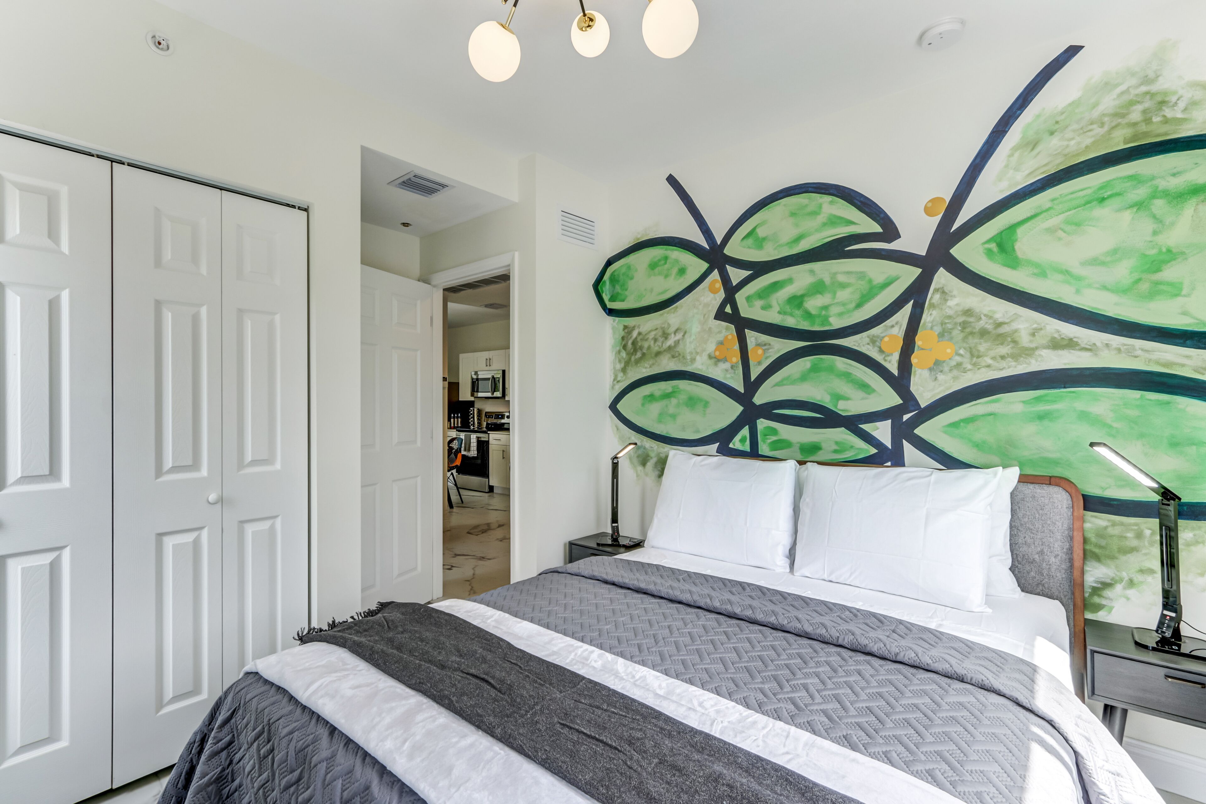 A neatly made bed with a gray and white striped bedspread, a modern headboard with a green abstract wall art, and a white wardrobe.