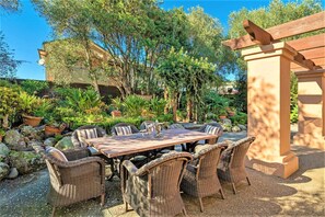 Un espace extérieur aménagé pour les repas, avec une table en bois et des chaises en osier, entouré de plantes en pot et d’arbres.