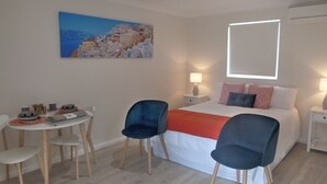 Ein Hotelzimmer mit einem Bett, zwei Stühlen, einem kleinen Tisch mit Schüssel und Tellern sowie einem Gemälde eines Küstdorfes.
