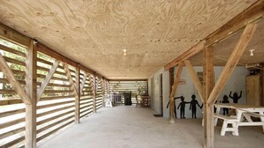 Ein überdachter Außenbereich mit Holzbalken und einem Betonboden, mit einer schlichten Bank und einer Wand mit Silhouetten von Personen.